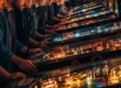 Fotografia de jogadores concentrados durante uma partida de torneio de pinball, com várias máquinas iluminadas ao fundo.